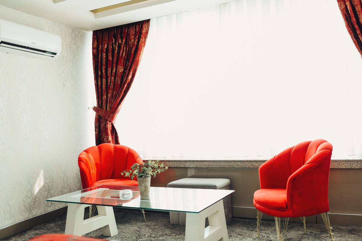 A room with a glass-top table, two red chairs, a small white stool, a vase with flowers, and a window with curtains.