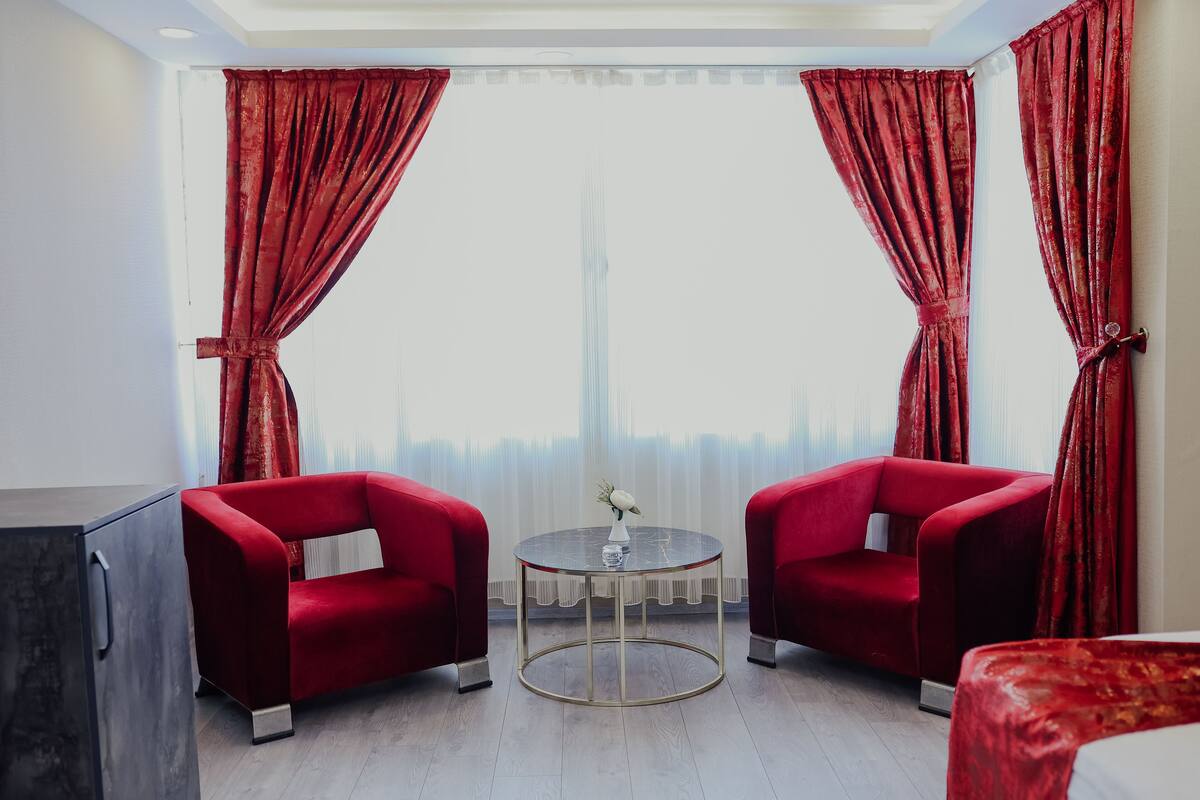 A hotel room with two red armchairs, a round glass table, and a view of a tall building.