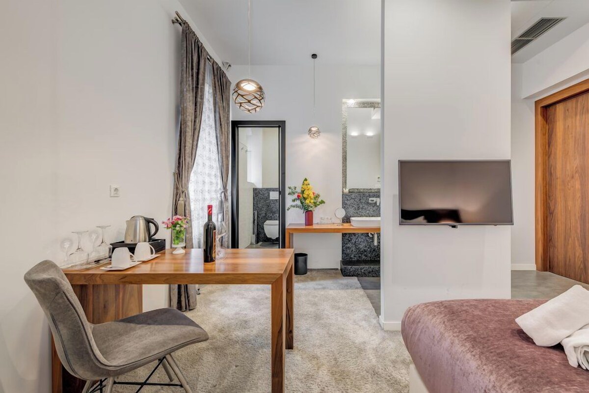 A modern hotel room with a wooden dining table, a gray upholstered chair, a pink bed, a TV mounted on the wall, and a bathroom with a mirror and sink.