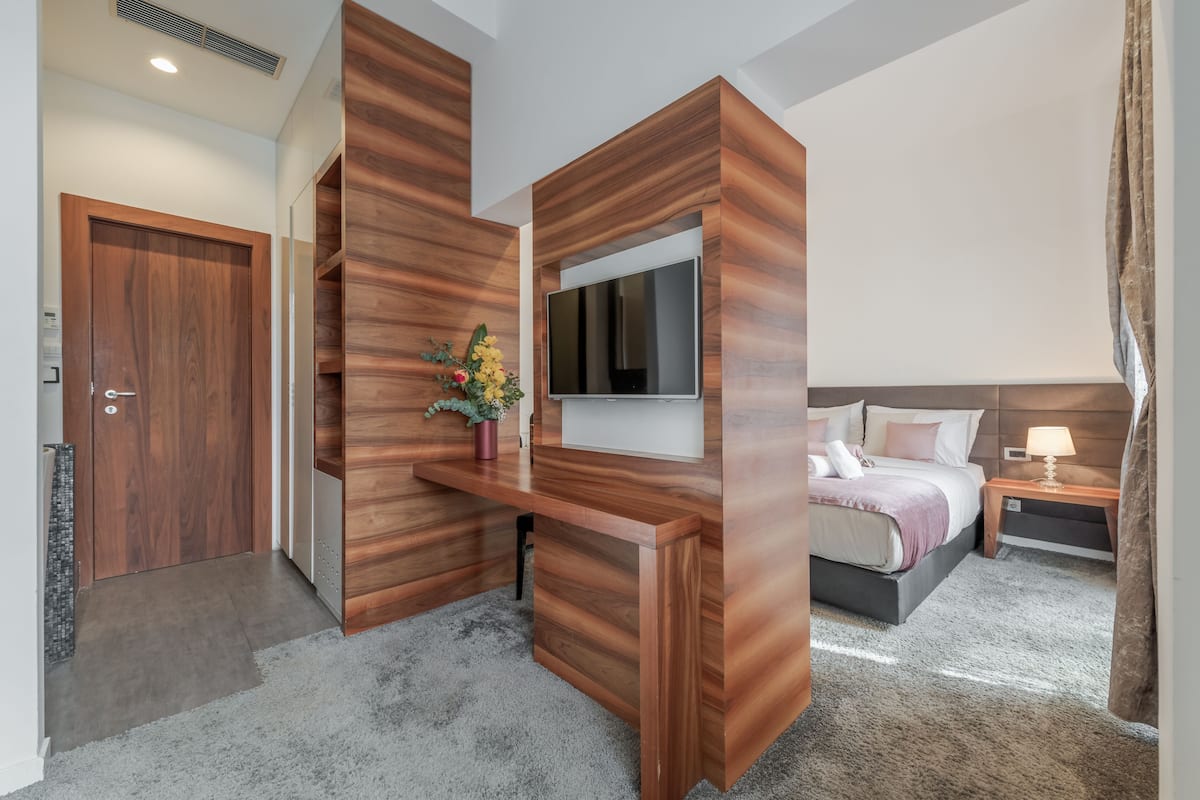 Una habitación de hotel moderna con un escritorio de madera, un televisor de pantalla plana, una cama con cabecera y una mesita de noche con lámpara.