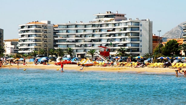 Un hotel frente a la playa con varios balcones, zona de piscina y sombrillas.