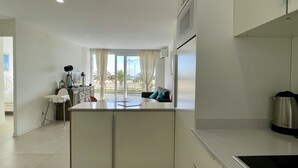 Una cocina moderna con una isla central, muebles blancos y vistas a palmeras a través de grandes ventanales.