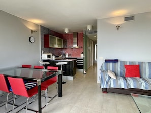 Eine moderne Küche mit Mikrowelle, Herd und einer roten, gefliesten Spritzschutzwand. Es gibt einen Essbereich mit roten Stühlen und einem Glastisch. Im Wohnzimmer steht ein gestreiftes Sofa mit roten Kissen.
