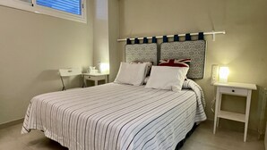 Une chambre à coucher comprenant un lit, deux tables de chevet, une commode, une fenêtre avec stores et un luminaire fixé au mur.
