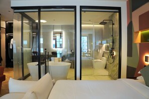 A modern hotel room with a large bed, a sitting area with a chair, a glass-enclosed shower area, and a visible bathroom with a bathtub. - Hotel Sapin (Bangkok)