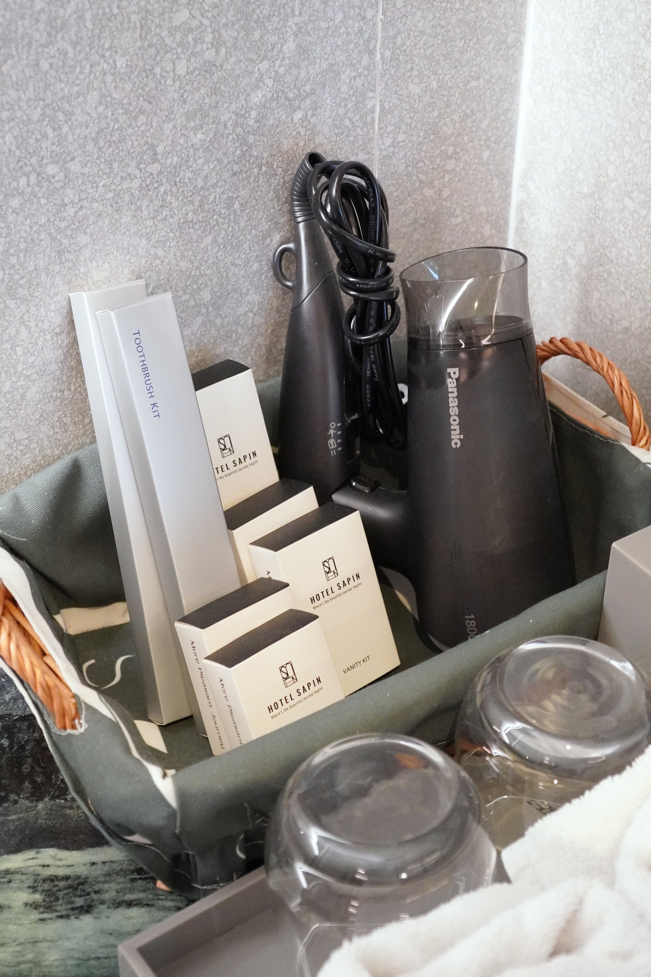 A basket containing toiletries including a toothbrush kit, a hairdryer, a Panasonic water bottle, and a glass.