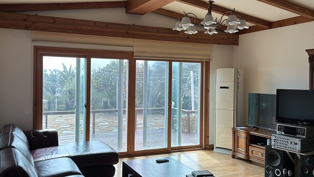 A spacious living room with a wooden ceiling, large sliding glass doors, a leather sofa, a coffee table, and a TV stand with speakers. - Betta's Homestay (Jeju City)