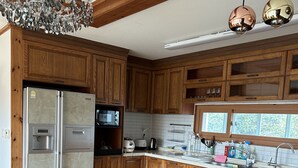 A kitchen with wooden cabinets, a stainless steel refrigerator, a microwave, a sink, and a wooden bench. - Betta's Homestay (Jeju City)