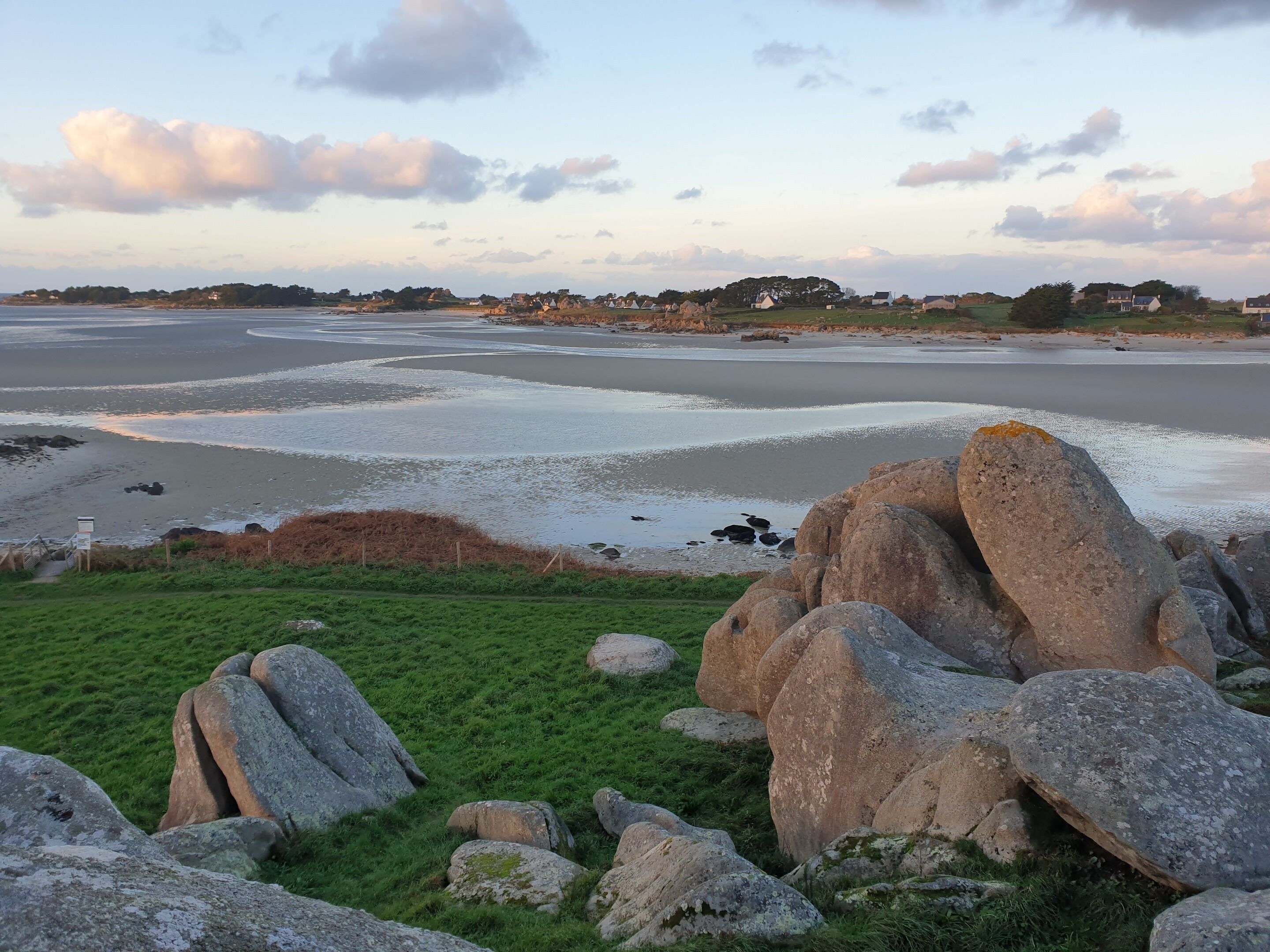 A coastal landscape with large boulders, grassy areas, and a beach with gentle waves.