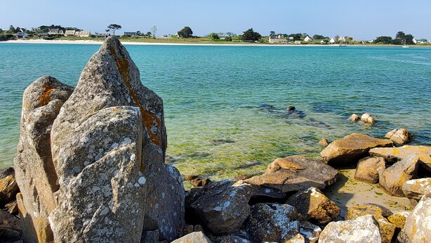 Paisaje costero con grandes rocas en primer plano y una vista despejada de la playa y las casas al fondo.