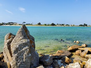 A coastal scene with large rocks in the foreground and a clear view of the beach and houses in the background.