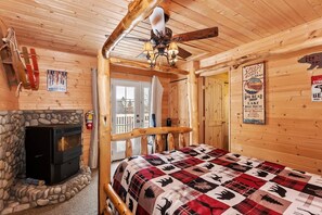 A cozy cabin interior with a stone fireplace, a bed with a patterned quilt, wooden walls, and a ceiling fan. - Pine Haven By Destination Big Bear: Close to the Village, Snow Summit and Marina's (Big Bear Lake)