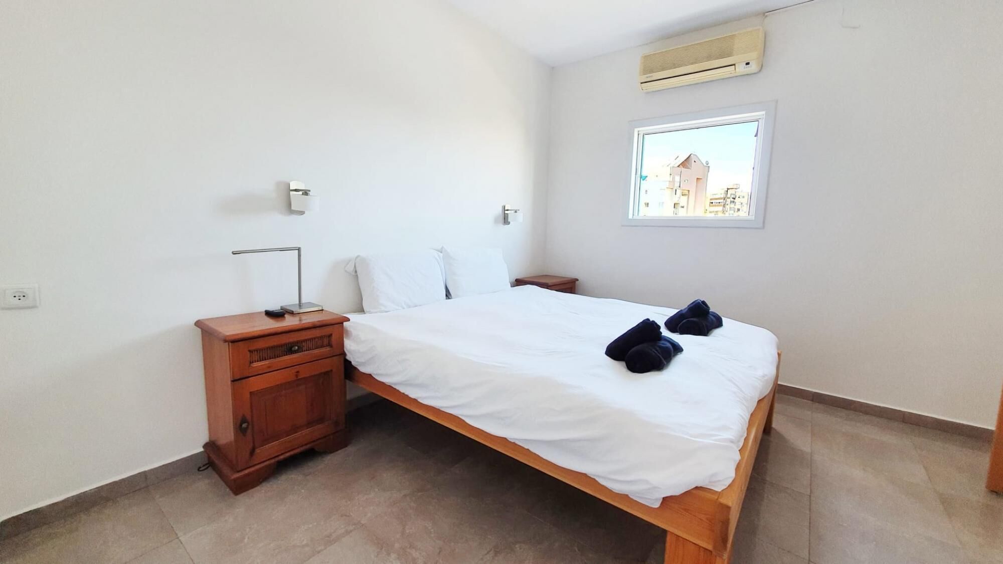 A bedroom with a wooden bed, white linens, a nightstand, a lamp, and a window with a view of buildings.