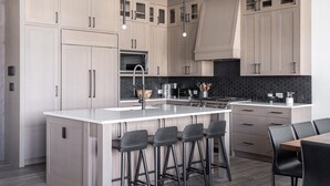 Una cocina moderna con armarios de madera clara, una isla central con taburetes de bar, un salpicadero de azulejos negros y electrodomésticos de acero inoxidable.