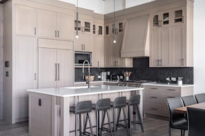 Een moderne keuken met lichte houten kastjes, een centraal eiland met barkrukken, een zwart betegelde spatwand en roestvrijstalen apparatuur.