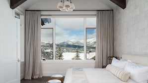 Un dormitorio con un amplio ventanal que ofrece vistas a la montaña, una cama con ropa de cama blanca y una lámpara de araña moderna.