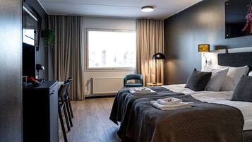 A hotel room with a bed, a desk, a chair, a radiator, a window with curtains, and a lamp.