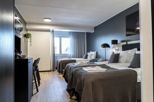 A modern hotel room with a large bed, a desk, and a chair. There is a window with curtains, a plant, and a lamp on the bedside table.