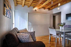 A modern living space with a wooden ceiling, a sofa with patterned cushions, a dining area with a table and chairs, and a kitchen with a built-in appliance.