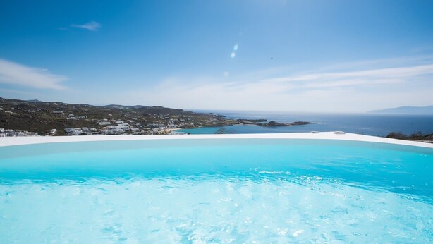 Ein klarer, blauer Infinity-Pool mit Blick auf eine Küstenlandschaft und ferne Berge.