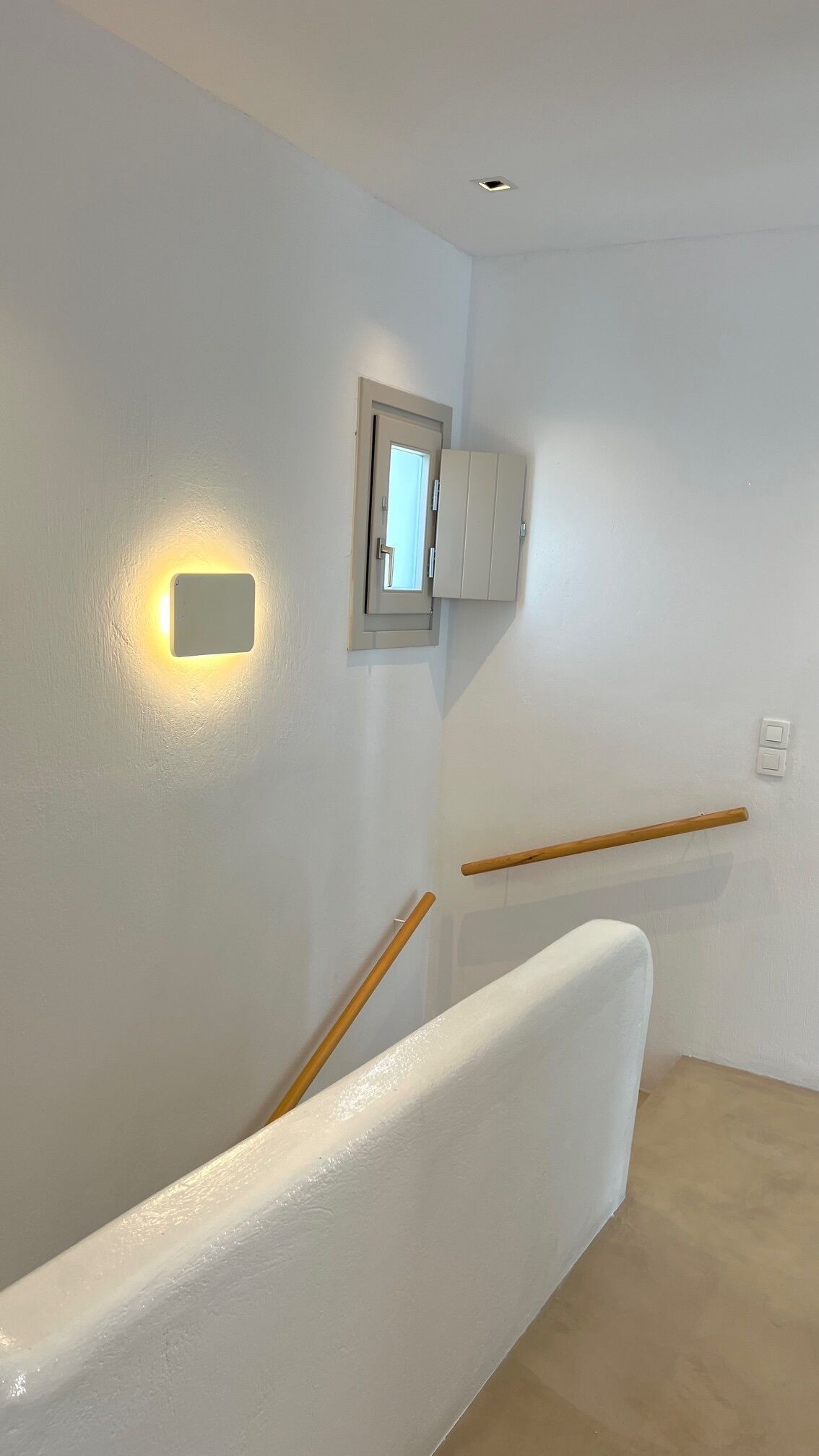 Un intérieur épuré, aux murs blancs, agrémenté d’une main courante en bois, d’un luminaire encastré au plafond et d’une petite fenêtre située au-dessus d’une porte.