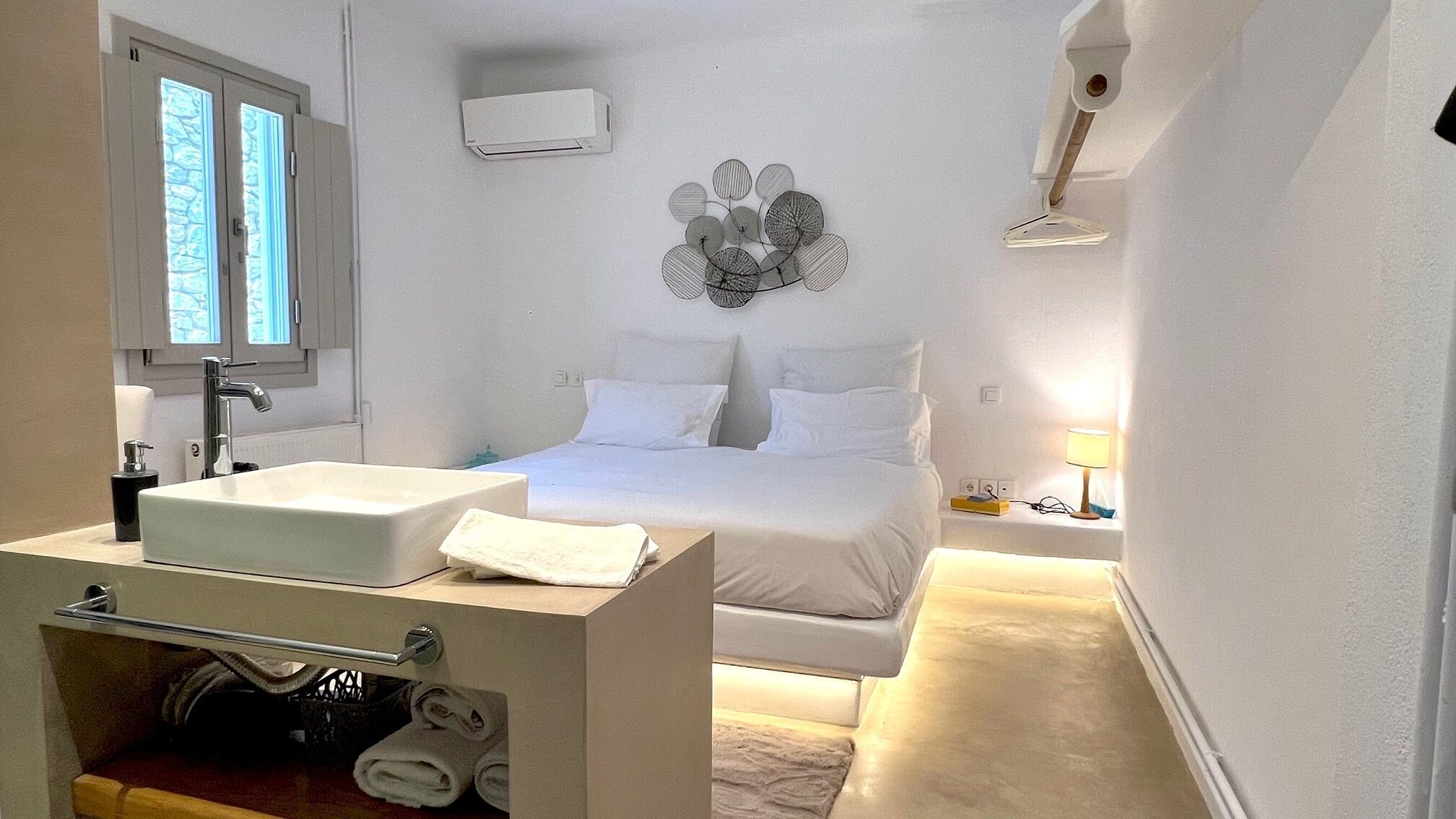 Une chambre à coucher comprenant un lit, une table de chevet avec une lampe, un climatiseur fixé au mur, et une salle de bain avec un lavabo et un porte-serviettes.