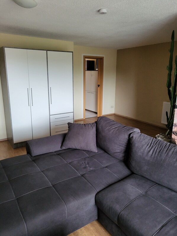 A living room with a grey sectional sofa, a white wardrobe, and a door leading to a kitchen.