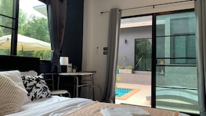 Ett hotellrum med en säng, ett litet bord med en lampa, utsikt över en swimmingpool och en skjutdörr i glas.