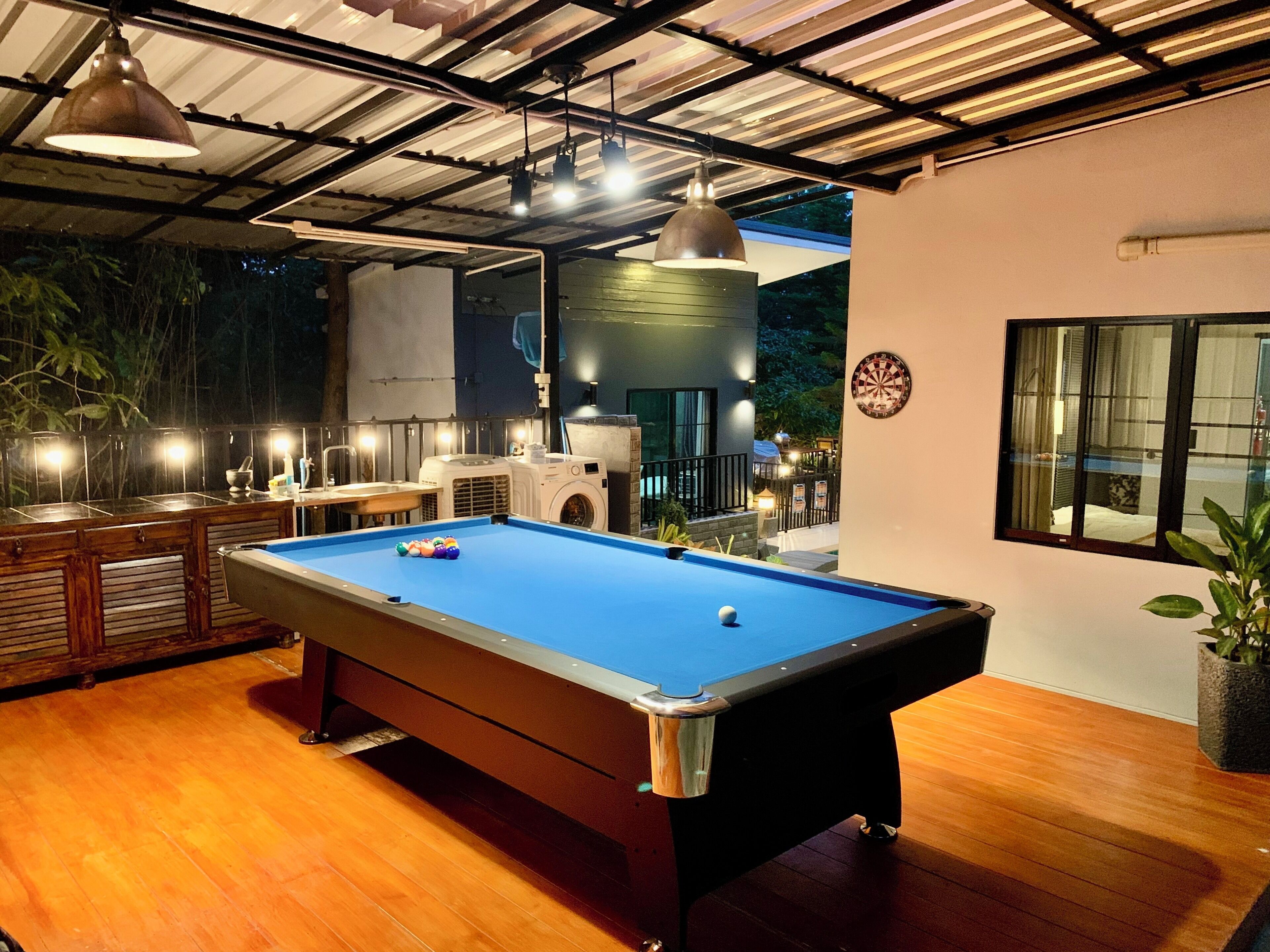 A pool table in a room with a high ceiling and wooden flooring. There are ceiling lights and a window with a view of the outside.