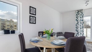 A dining area with a round table, dark chairs, and a vase with flowers. There are framed pictures on the wall and a window with a view of buildings. - Comfortable Apartment in the Center | Balcony and Parking (Kolobrzeg)