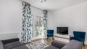 A modern living room with grey sofas, a flat-screen TV on a white console, and a large window with patterned curtains. - Comfortable Apartment in the Center | Balcony and Parking (Kolobrzeg)