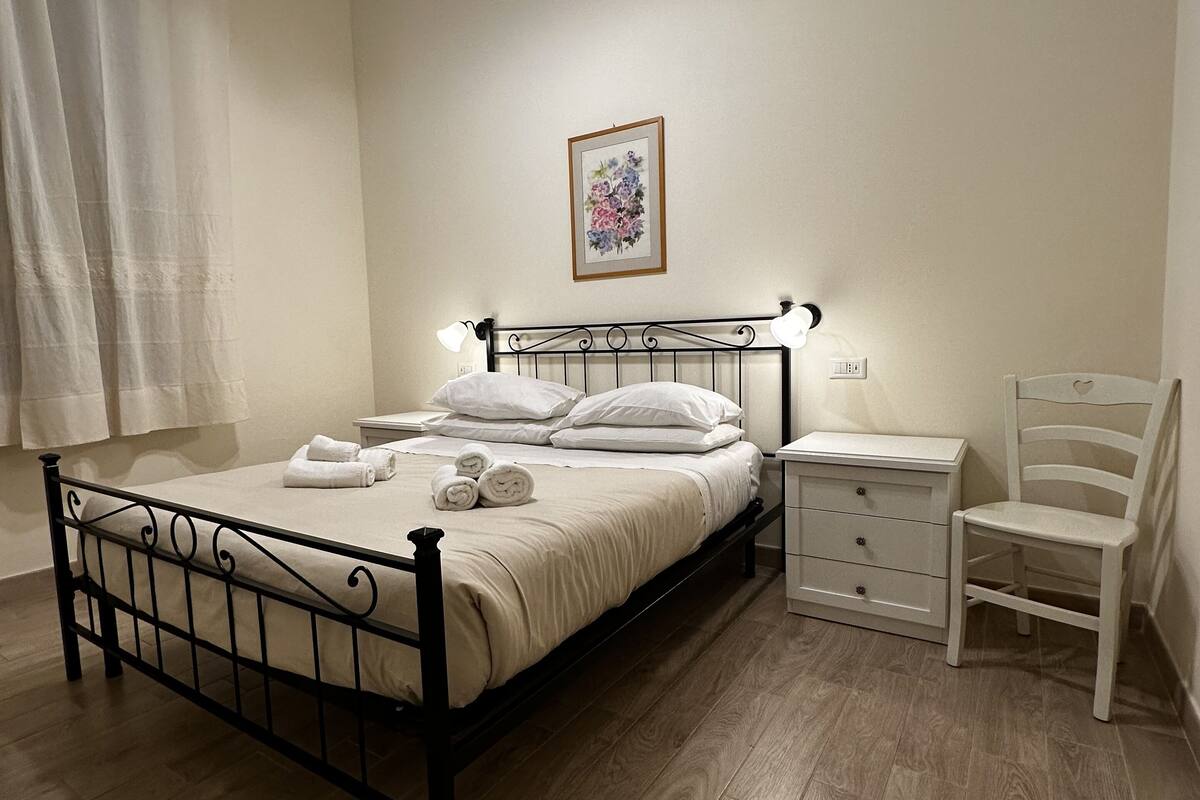 A neatly made bed with white linens, a bedside table with a lamp, a chair, and a wall-mounted picture.