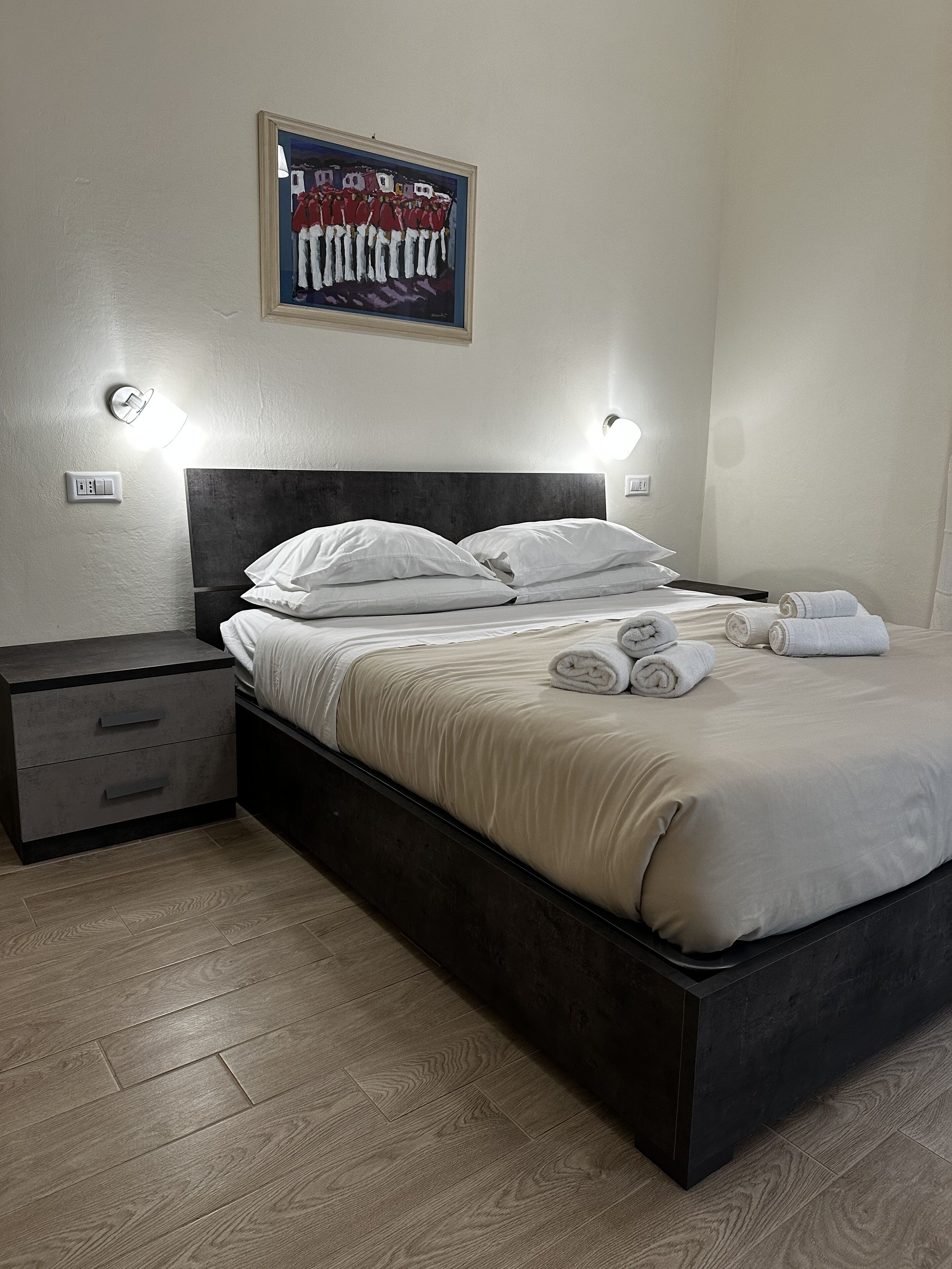 Ein ordentlich bezogenes Bett mit weißen Leinen und aufgerollten Handtüchern, ein dunkles Kopfteil und ein an der Wand hängendes Bild.