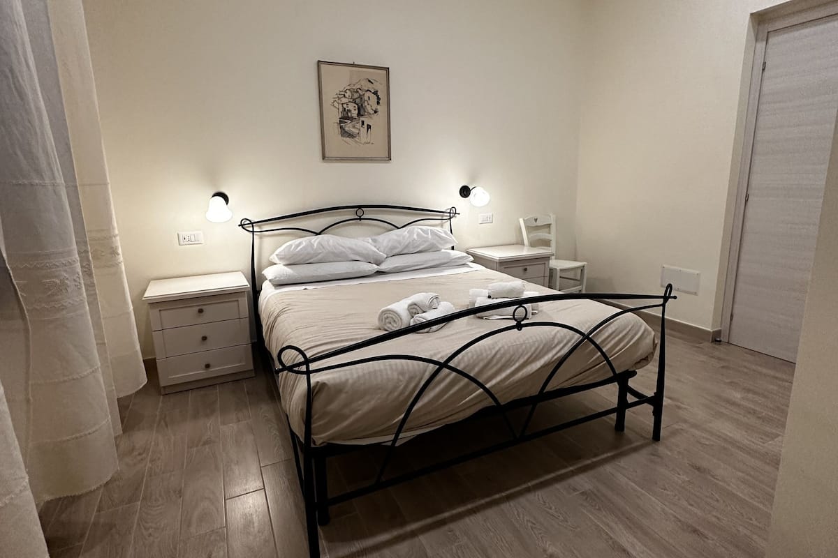A bedroom with a bed, bedside tables, a nightstand, a wall-mounted lamp, and a framed picture.