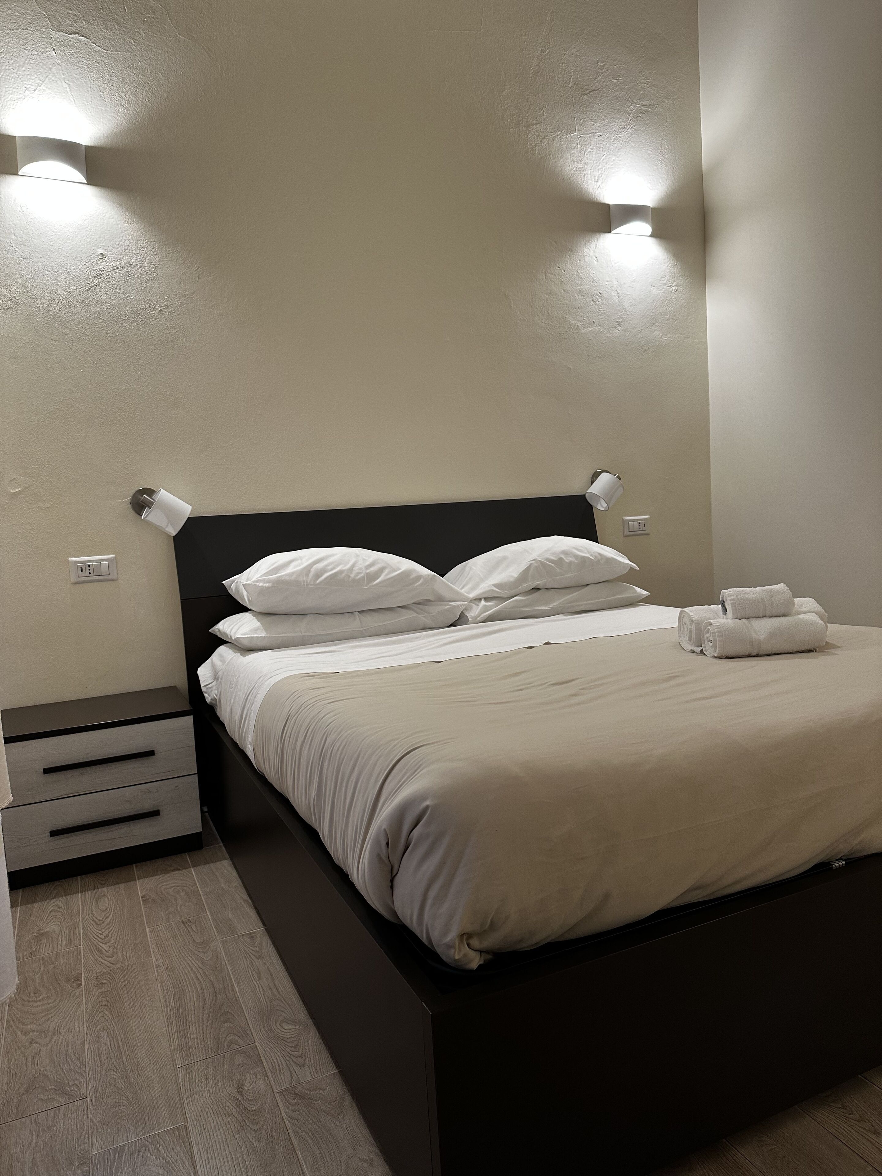 A neatly made bed with white linens, a black headboard, and two wall-mounted lights.