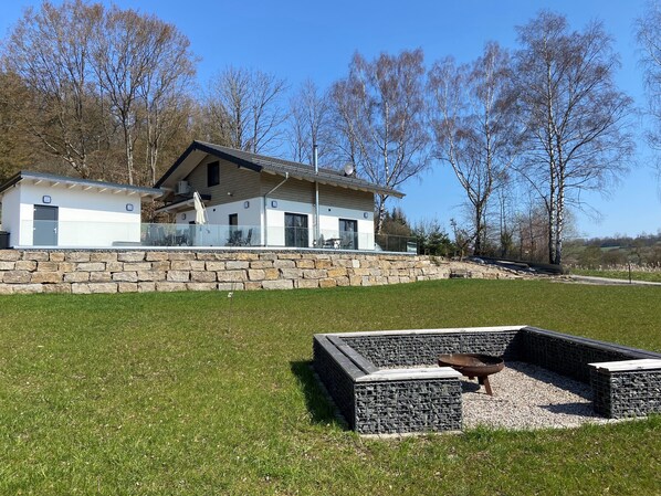 A modern house with a stone retaining wall, a fire pit, and a grassy yard. - Vacation Home in the Rhön (Kaltennordheim)