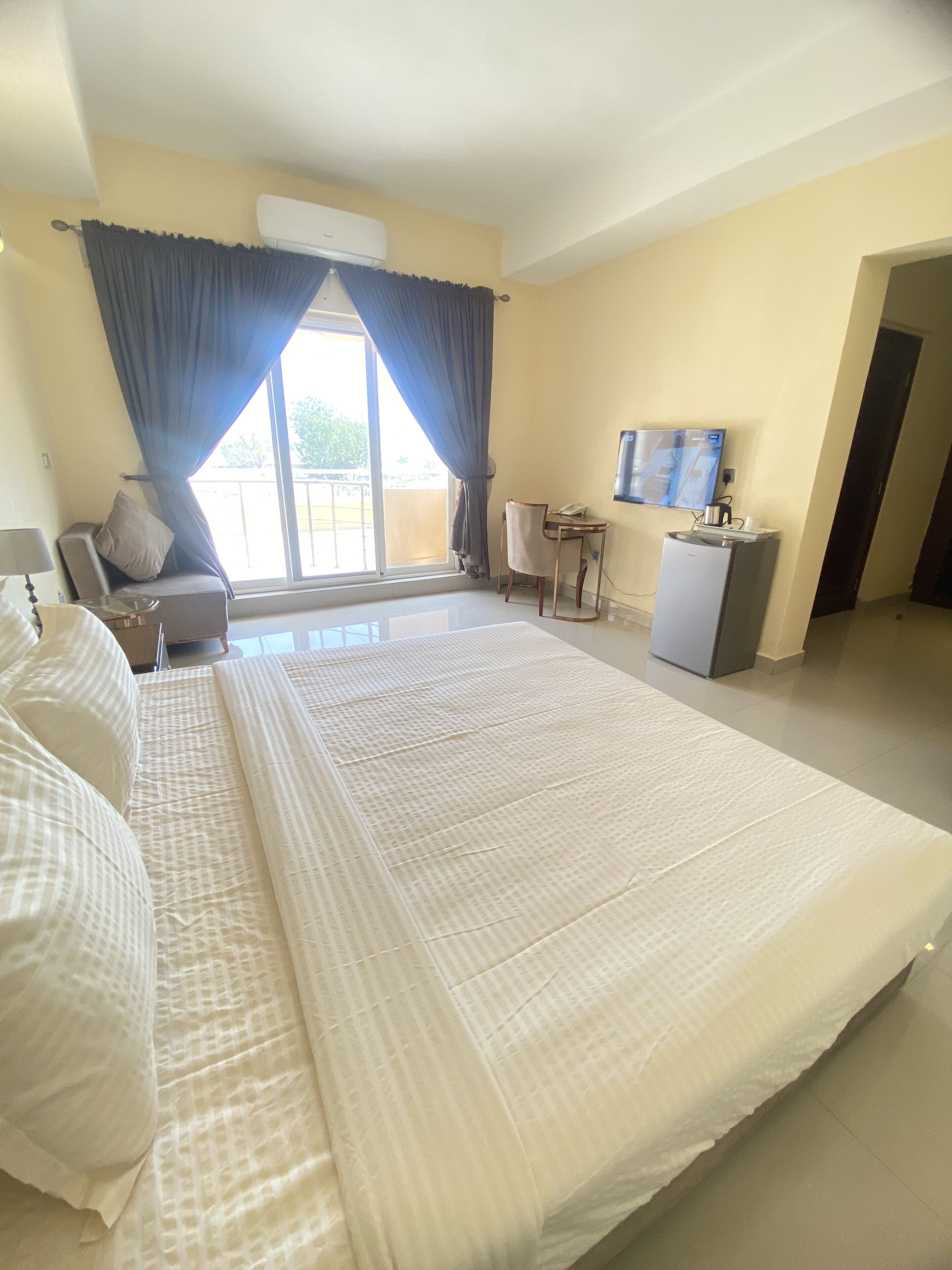A hotel room with a large bed, a desk, a chair, a TV, and a balcony with curtains.