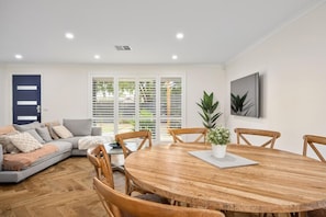 A modern living room with a wooden dining table, grey sofa, and a large window with blinds.