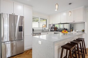 A modern kitchen with white cabinetry, a stainless steel refrigerator, a large island with stools, and pendant lighting. - Secret Getaway, Capel Sound (Capel Sound)