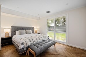 A bedroom with a large bed, a bench, a nightstand, and a window with blinds.