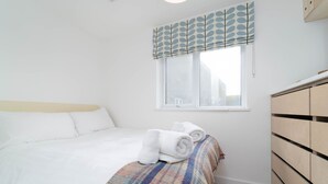 Een slaapkamer met een bed, een raam met een stoffen gordijn, een houten dressoir en een geruite deken.