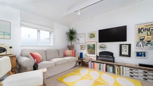 Een woonkamer met een bank, een salontafel, een geel vloerkleed, een boekenkast en een flatscreen televisie aan de muur.