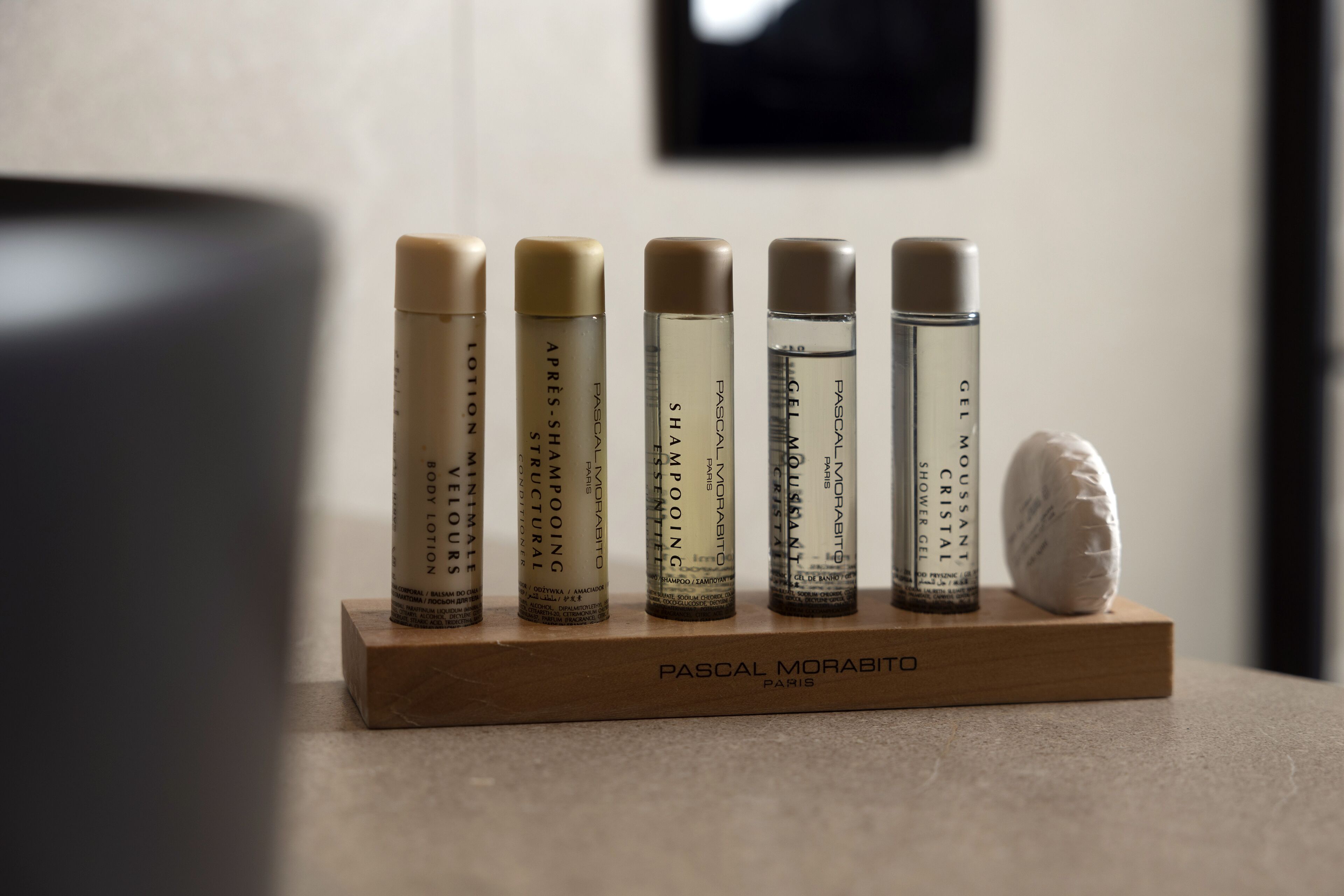 Un présentoir en bois contenant six bouteilles de lotion pour le corps de Pascal Morabito et un petit objet blanc.