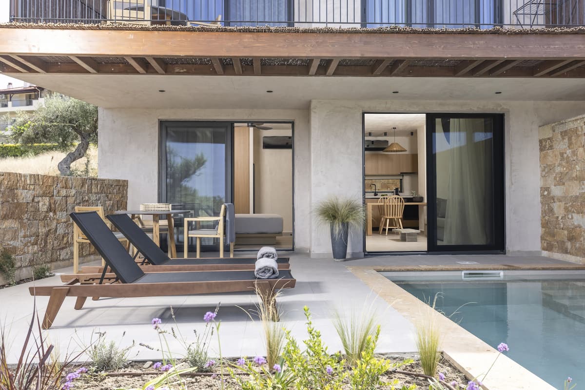 A modern outdoor pool area with lounge chairs, a dining table, and a kitchen visible through glass doors.