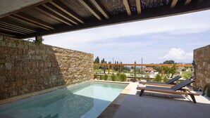 Piscina con paredes de piedra y sillas de madera en la terraza, con vistas a una zona residencial.