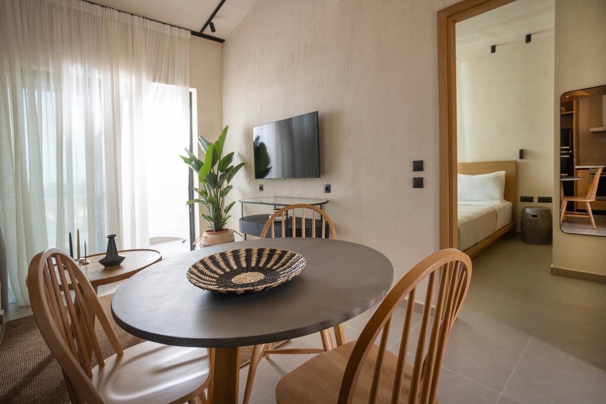 A modern hotel room with a dining area, a flat-screen TV mounted on the wall, and a view into the adjacent bedroom.