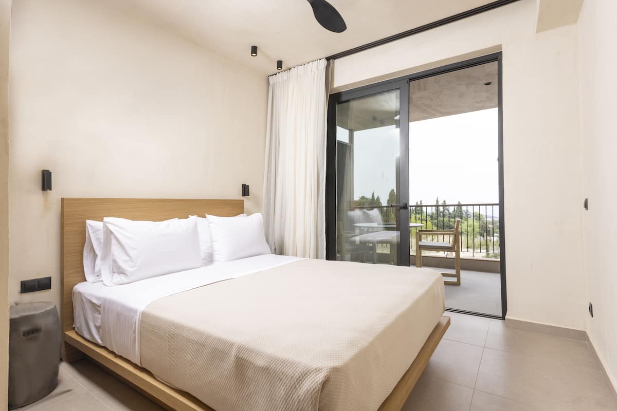 A hotel room with a bed, a small trash bin, a ceiling fan, and a balcony with a view.