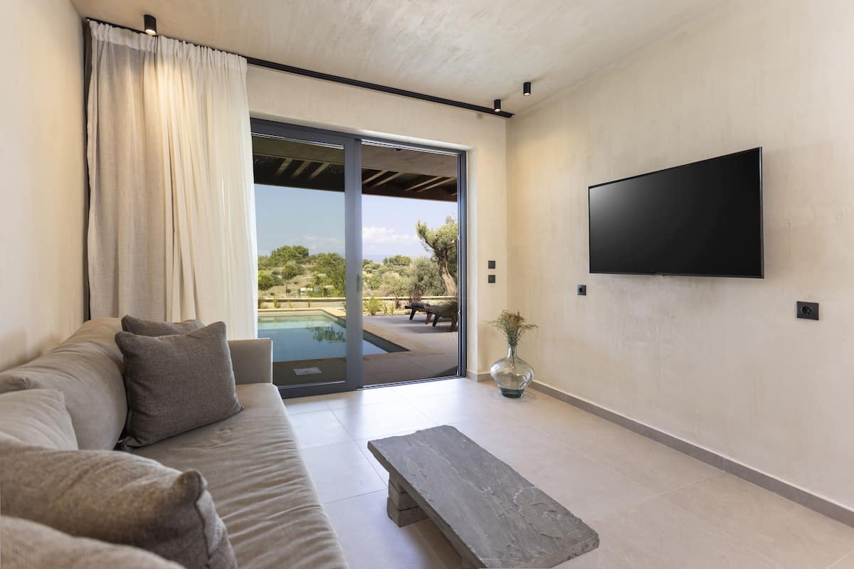 A modern living room with a sofa, a coffee table, a flat-screen TV, and a sliding glass door leading to an outdoor area with a pool.