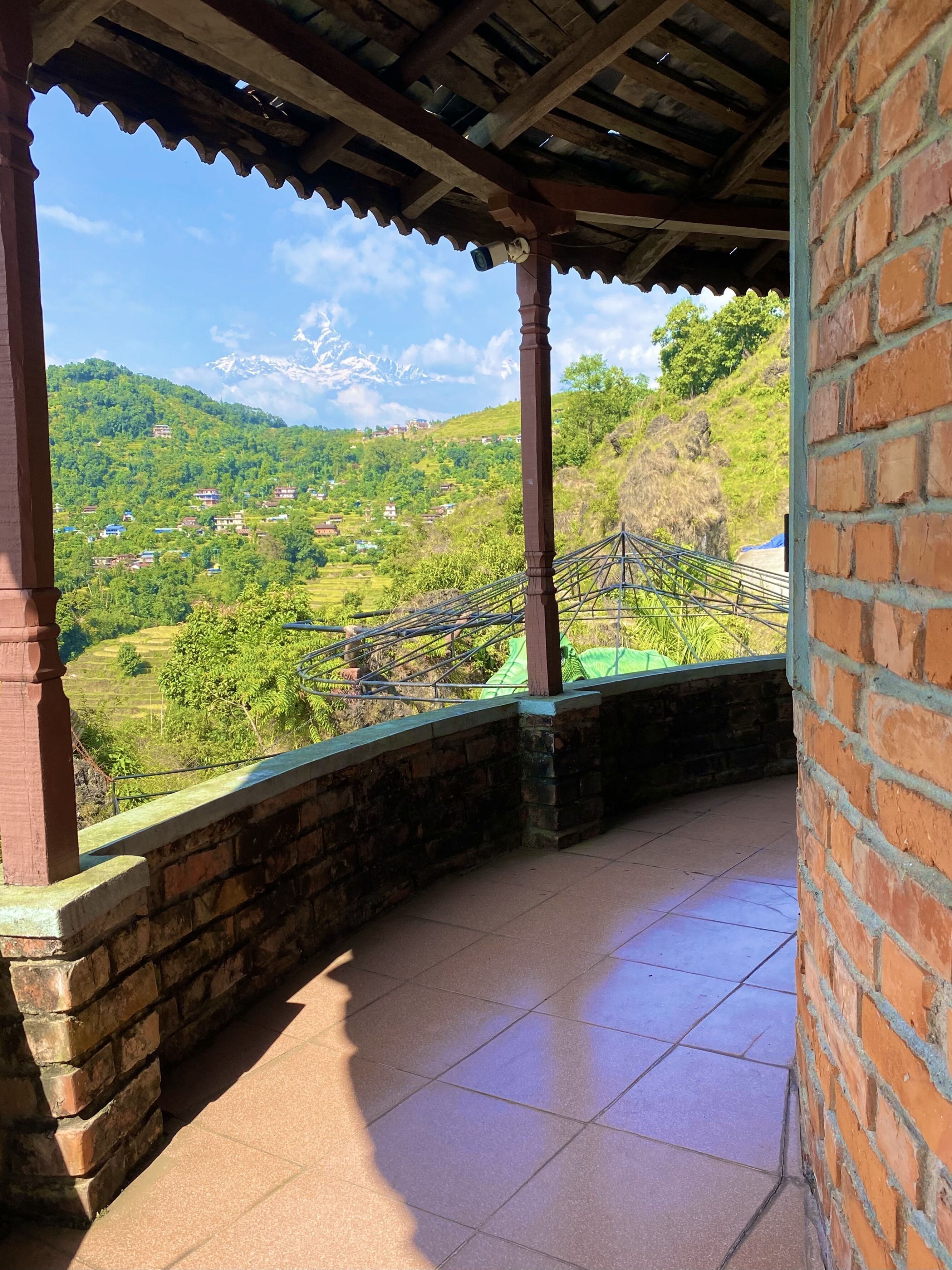 A covered balcony with a view of a green valley and distant mountains.