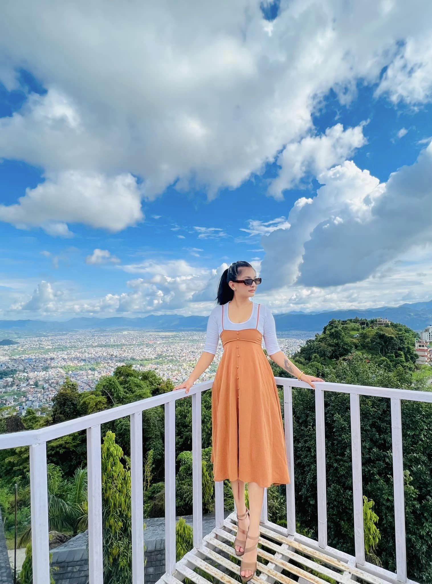 A person standing on a white metal railing overlooking a cityscape with mountains in the background.